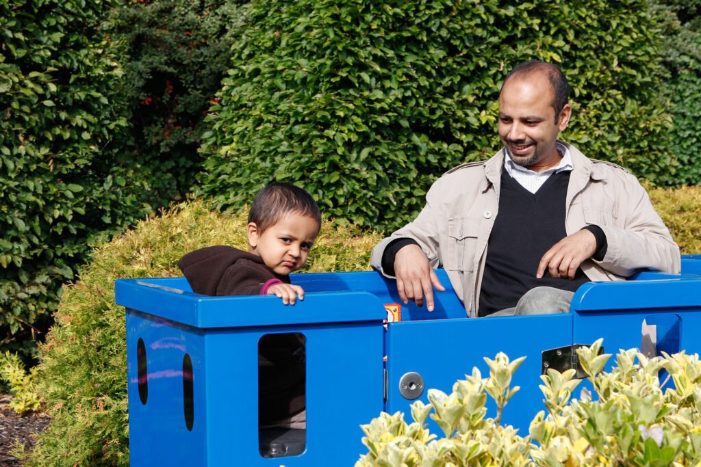 A young boy sits in a blue play structure, looking curious, while his father smiles warmly at him, surrounded by lush green foliage in an outdoor setting.